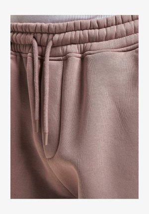 Heledad roosa dressipüksid pehmest kangast, elastse vöökoha ja reguleeritavate paeltega, millel on plastikkatted. Küljepocketi detail.