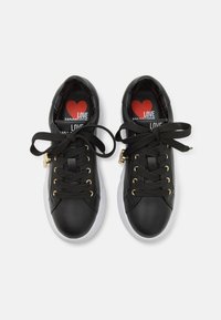Black athletic shoes with a smooth leather finish, gold eyelets, black laces, and white rubber soles. Interior features a red heart graphic.