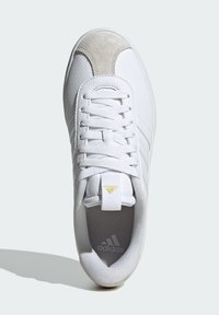 White athletic shoe featuring a textured leather upper, suede toe accent, six lace eyelets, and a gold logo on the tongue and insole.