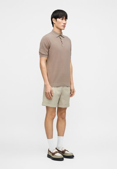 Φωτεινό καφέ πλεκτό πόλο με κοντά μανίκια, συνδυασμένο με μπεζ σορτς. Φοράε ι άσπρες κάλτσες και casual παπούτσια με σκούρα σόλα.