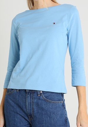Kobieta ubrana w jasnoniebieski, długorękawowy top z małym logo na piersi, w połączeniu z wysokim stanem niebieskich dżinsów.