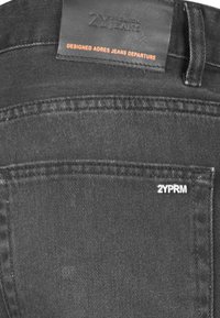 Crne traperice od denima s kožnom oznakom na kojoj piše "2Y PREMIUM JEANS" i detaljima šivanih džepova. Sadrži bijeli "2YPRM" brendiran tiskan na džepu.