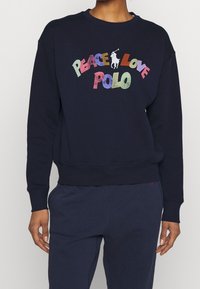 Námořnická modrá mikina s žebrovanými manžetami a lemem, s barevným nápisem "PEACE LOVE POLO" a bílým logem koně. Neformální design.