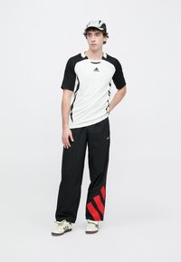 Športový outfit pozostávajúci z bielo-čierneho trička s krátkym rukávom a logom Adidas, v kombinácii s čiernymi nohavicami s červenými pruhmi, doplnený čiapkou.