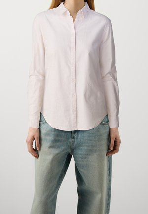 Kvinde iført en lyserød, langærmet skjorte med knapper, der ikke er stoppet ned i, over lyseblå, falmede jeans, stående foran en ensfarvet baggrund.