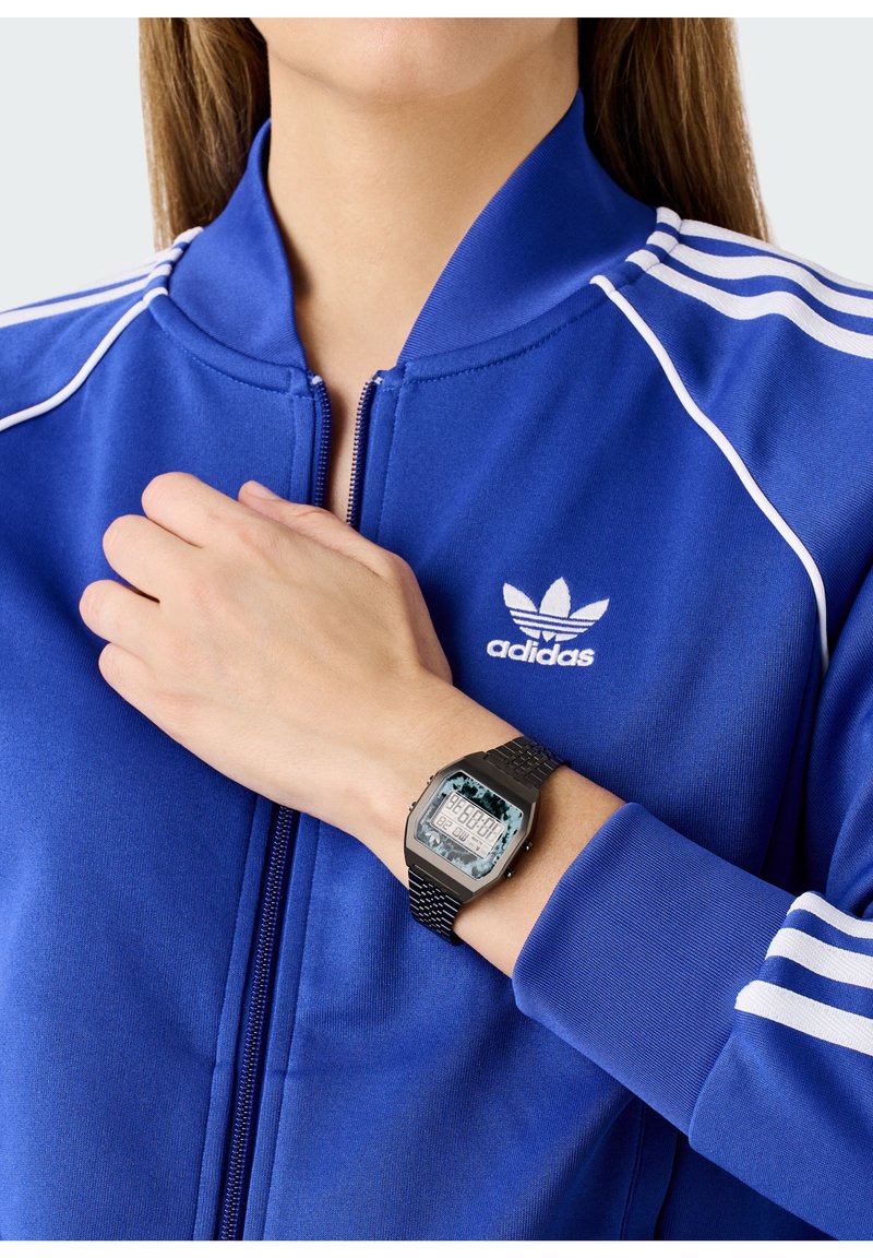 Veste de survêtement zippée bleue avec des rayures blanches ; montre numérique noire au poignet, avec un cadran carré et un bracelet texturé. Logo Adidas visible.