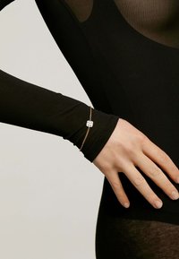 Pulsera de cadena de oro con una piedra rectangular transparente, que se lleva sobre una camiseta de manga larga negra; textura suave y diseño minimalista.