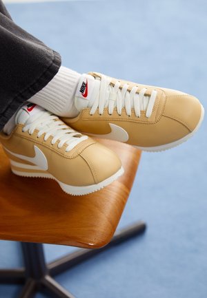 Personne portant des baskets Nike Cortez couleur tan et des chaussettes blanches, les pieds reposant sur une chaise en bois sur un fond bleu.