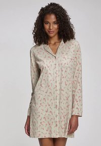 Beige pysjamaskjorte med lange ermer, mønstret med røde sukkertøystokker. Har en fremre lomme og knappelukking. Glatt stofftekstur.