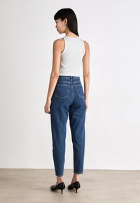 Grijze mouwloze top gecombineerd met high-waisted blauwe denim jeans. De jeans hebben achterzakken en een taps toelopend ontwerp. Zwarte hakken maken de outfit compleet.