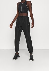 Černý sportovní outfit sestávající z přiléhavého krátkého topu a volných kalhot, obojí s šedým prošíváním a elastickými manžetami, doplněný černými teniskami.