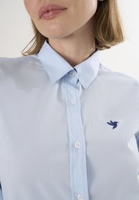 Lysblå skjorte med knapper i glat stof, der har en lille navy fuglebroderi på venstre side og et klassisk kravedesign.