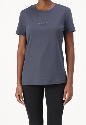 Woman wearing a fitted short-sleeve navy blue Calvin Klein Jeans t-shirt and black pants, standing against a plain white background.