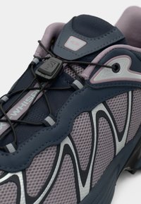 Zapatillas deportivas con parte superior de malla gris, acentos morados y suela de goma moldeada. Presentan cordones elásticos y un detalle de logotipo prominente.