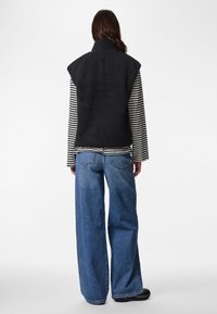 Crna prsluk od šerpe s visokim ovratnikom, nošen preko crno-bijele prugaste košulje s dugim rukavima, uparene sa širokim plavim trapericama.