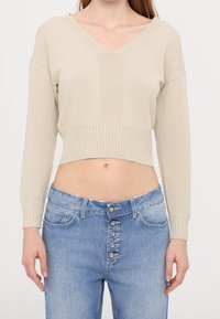 Suéter corto color beige con escote en V, detalle de costillas verticales y mangas largas. Combina con jeans azul claro de talle alto y cierre de botones.