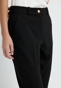 Close-up van een persoon die zwarte op maat gemaakte broek met een gouden knoop draagt, een witte top en met de linkerhand vlakbij de heup rust.