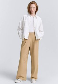 Weiße, leichte Jacke mit Kragen und vorderen Knöpfen, kombiniert mit beigen, weit geschnittenen Hosen und weißen Turnschuhen. Weiche Stofftextur.