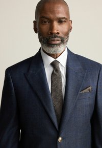 Blazer à carreaux bleu marine avec un bouton en or, chemise blanche, cravate brune texturée et pochette brune.