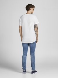 Weißes Kurzarm-T-Shirt, lässiger Schnitt, kombiniert mit hellblauen Slim-Fit-Jeans und marineblauen Sneakern mit weißen Sohlen. Sichtbare Tattoos am Arm.