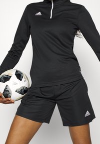 Personne en tenue de sport Adidas noire tenant un ballon de football Adidas blanc avec des motifs pixellisés noirs sur un fond blanc uni.