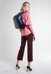 Mochila de cuero navy con acentos turquesa, de forma rectangular, textura suave, llevada por un modelo con un suéter rosa y pantalones burdeos.