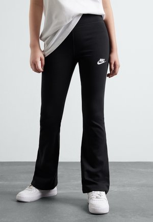 Osoba ubrana w czarne, rozszerzane legginsy Nike z białym logo i białe trampki, stoi na szarej podłodze na jednolitym tle.