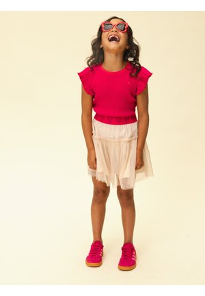 Meisje met rode zonnebril, rood shirt met geruite mouwen, beige rok en rode sneakers, staand en lachend terwijl ze omhoog kijkt.