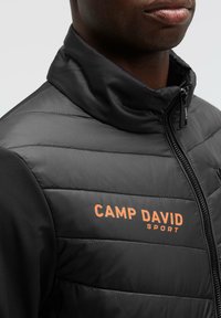 Chaqueta negra aislante con textura acolchada y cuello alto. Presenta un logo naranja "CAMP DAVID SPORT" en el pecho y cierre de cremallera.