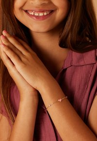 Gouden armband met roze hart en kraalaccenten, gestructureerde ketting, gedragen om de pols van een persoon met samengevoegde handen.