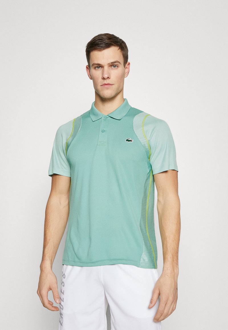 Polo vert avec manches courtes, présentant des panneaux latéraux en mesh, un col et un petit logo sur la poitrine. Texture lisse et coupe athlétique.