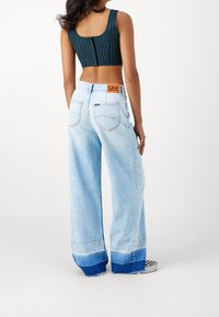 Sininvihreä raidallinen lyhyt yläosa yhdistettynä vaaleansinisiin leveälahkeisiin farkkuihin, joissa on rispaantunut helma ja takana Lee-merkki.