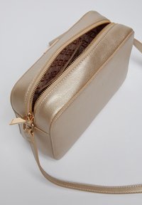 Beige texturovaná kožená crossbody taška se zlatým zipem, vnitřním vzorovaným látkovým podšívkou a úzkým popruhem na světlém pozadí.