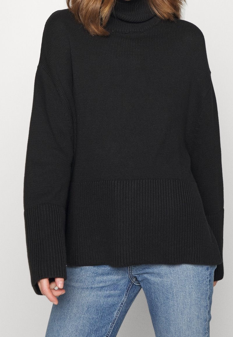 Pull à col roulé en tricot noir avec un design en côtes texturé, une silhouette oversize et des épaules tombantes, complété par des manches longues.
