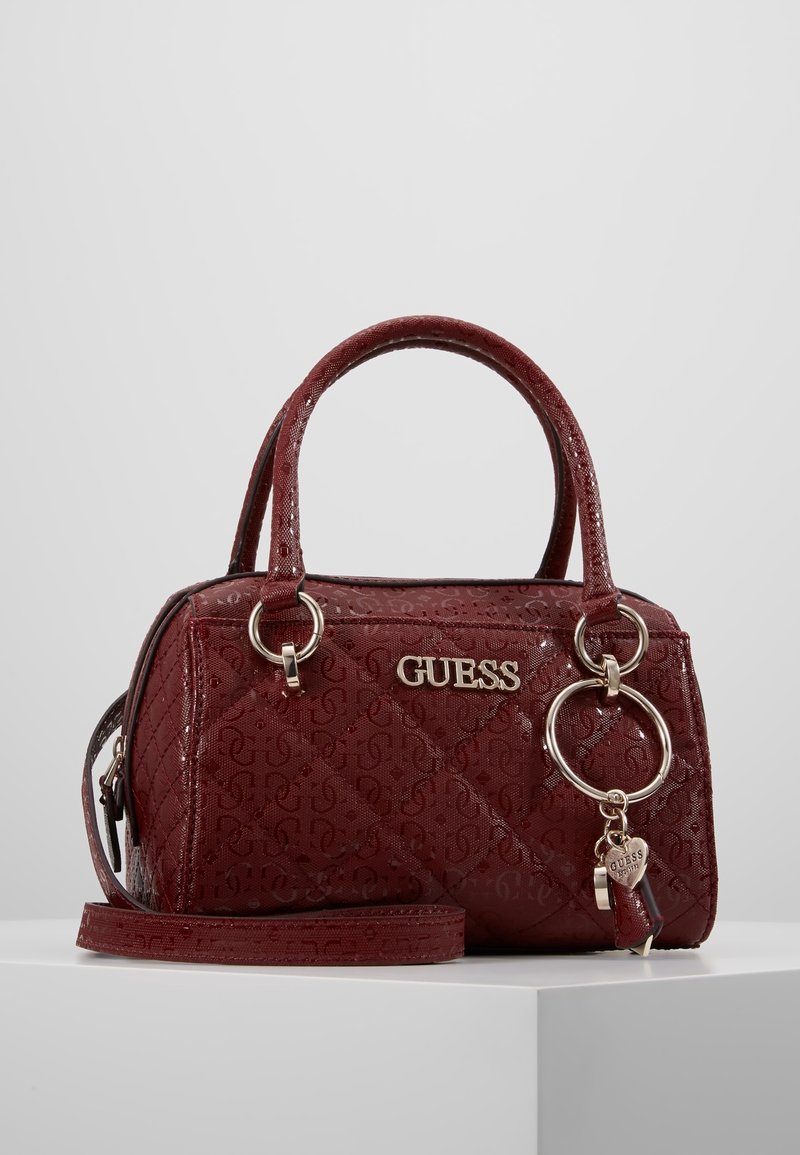 Sac à main Guess bordeaux avec motif matelassé, double poignée, bandoulière amovible et porte-clés rond avec breloque en forme de cœur sur fond blanc.
