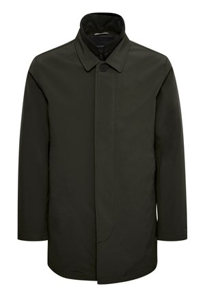 Homme portant un manteau noir, une chemise beige, un pantalon marron et des chaussures noires, debout avec une main dans la poche sur un fond uni.