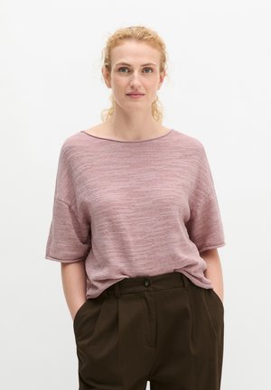 Vrouw met krullend blond haar, die een loszittend roze gebreid topje en donkerbruine hoge taillebroek draagt, handen in de zakken, staand tegen een effen achtergrond.
