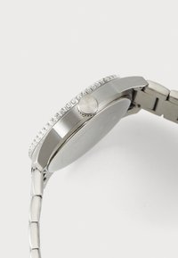 Montre en acier inoxydable avec un boîtier rond, une lunette cannelée, et un bracelet lisse et brossé. Elle possède une couronne sur le côté et une surface polie.