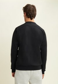 Uomo con felpa nera semplice e pantaloni tuta grigio chiaro che guarda verso il basso su uno sfondo neutro.