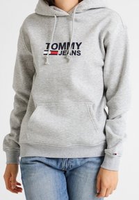 Felpa grigia con tasca a marsupio e cappuccio regolabile con coulisse. Presenta il logo "TOMMY JEANS" in blu navy e rosso sul petto. Polsini e orlo a costine.