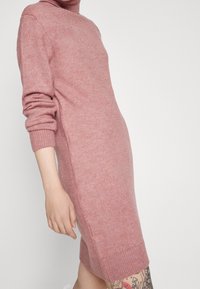 Abito lavorato a maglia in morbido tessuto rosa, caratterizzato da un colletto alto, maniche lunghe e polsini a coste. Forma aderente con un orlo di lunghezza media.