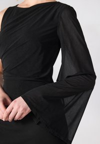 Svart klänning med en axel-design och en genomskinlig, utsvängd lång ärm. Tyget är slätt och figurnära, med subtil drapering som accentuerar.