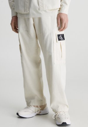 Mann trägt cremefarbene Cargohosen mit CK Jeans Logo-Patch und weißen Turnschuhen, steht vor einfarbigem Hintergrund.