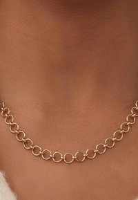 Collier chaîne en or avec des maillons circulaires entrelacés porté autour du cou d'une personne sur un fond neutre.