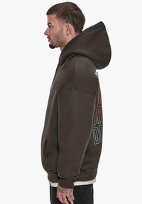 Brun hoodie med en stor hætte, sidelomme og farverig tekst trykt på ryggen. Har en ribbet kant og manchetter. Produkter i en bomuldsblanding.