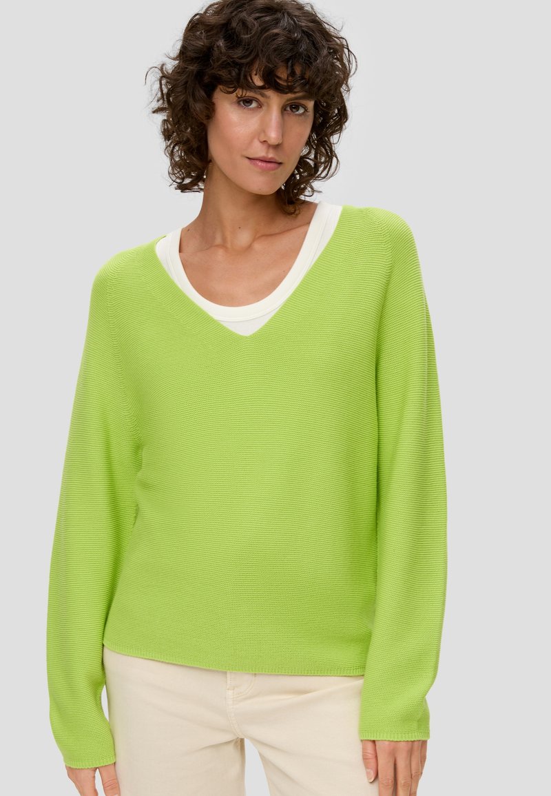 Jasnozielony, wełniany sweter z dekoltem w serek i długimi rękawami, noszony na białej koszulce z okrągłym dekoltem. Gładka faktura i luźny krój.