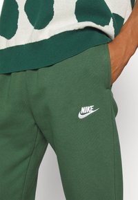 Zelené joggingové kalhoty vyrobené z bavlněného směsového materiálu. Mají bílé logo Nike a boční kapsy, s hladkou texturou a elastickým pasem.