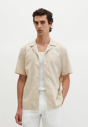 Jeune homme aux cheveux bouclés foncés portant une chemise beige à manches courtes boutonnée par-dessus un débardeur blanc et un pantalon blanc, debout devant un fond uni.