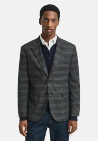 Grauer Karoblazer mit Reverskragen, zwei Knöpfen und vorderen Taschen. Über einem marineblauen Pullover getragen, kombiniert mit Jeans. GANT-Logo am Ärmel.