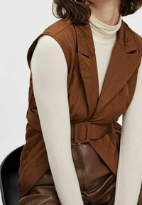 Braun gesteppte ärmellose Weste mit einem Gürtel, über einem cremefarbenen Rollkragenpullover getragen. Der Stoff wirkt weich und strukturiert und betont die Form des Outfits.